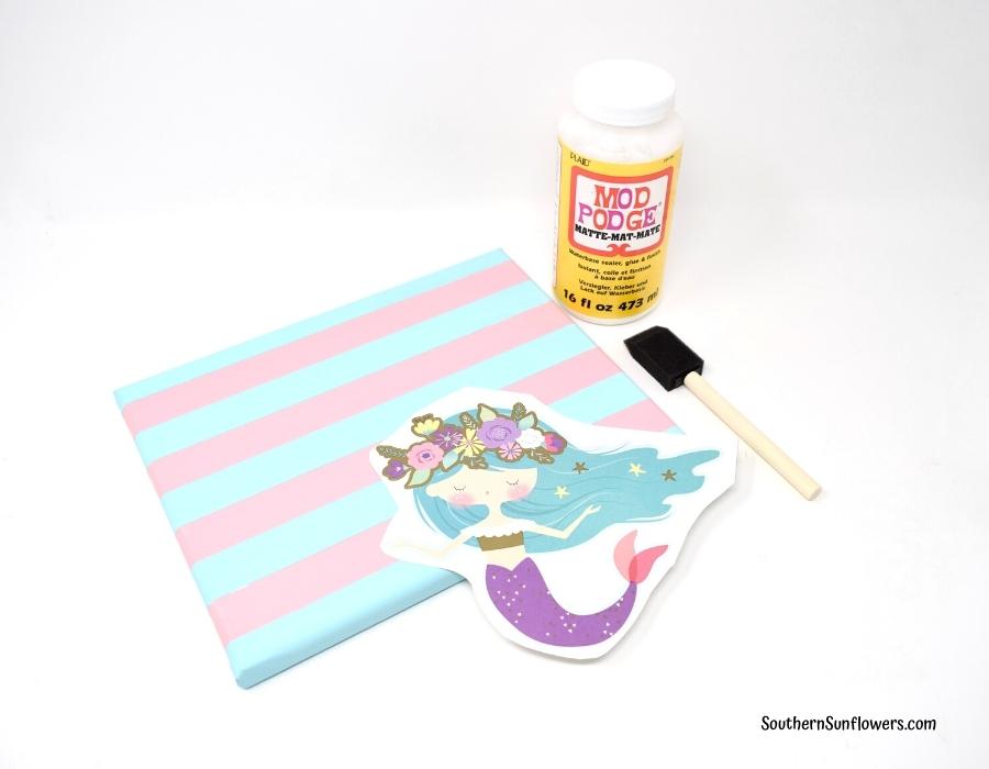 step for applying dollar tree stickers to canvas ~ mod podge bottle, brush, sticker, and painted canvas